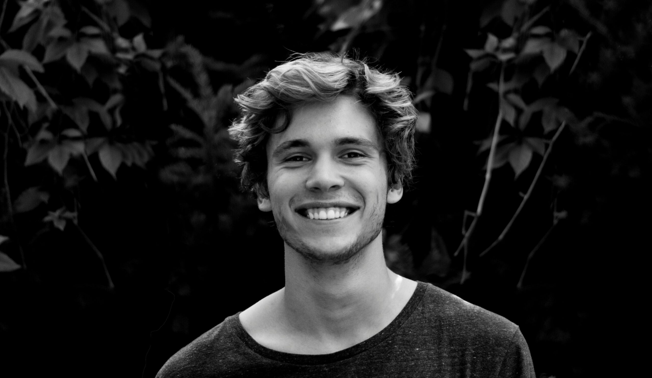Home A black and white portrait of a young man smiling outdoors with natural background.